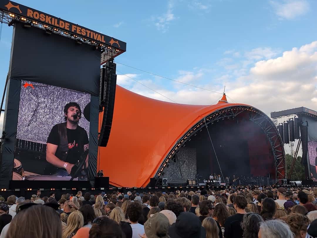 Wolf Alice toca en el escenario naranja del Roskilde Festival, con la cara de uno de sus músicos en la pantalla gigante y el público de espaldas, en julio de 2026 (foto de archivo). Foto: SkärFoto, CC BY 4.0, vía Wikimedia Commons
