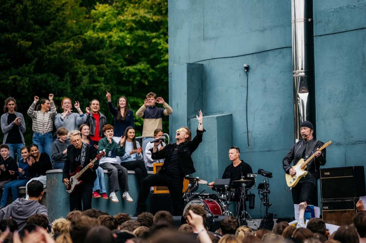 Foto de prensa oficial de U2 tocando en vivo en el patio del colegio Mount Temple de Dublín, el 25 de septiembre de 2026: Bono, con anteojos de sol amarillos y campera negra, canta con el brazo en alto en el centro; a la izquierda, Adam Clayton con un bajo rojo; atrás, Larry Mullen Jr. en la batería, y a la derecha, The Edge con gorro negro y guitarra clara, frente a una pared verde azulada, con alumnos sentados sobre un muro y otros aplaudiendo. Foto: Rich Fury