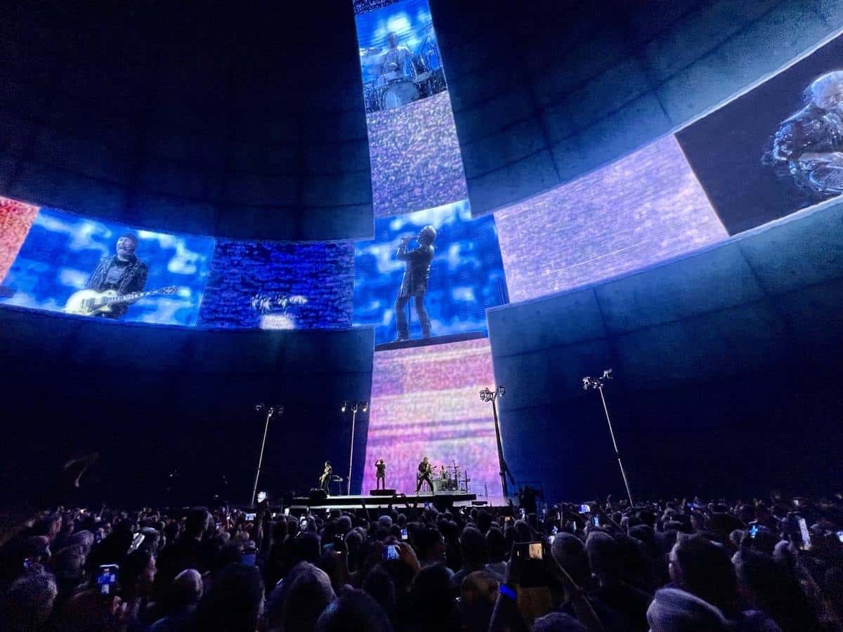 U2 en vivo en la Sphere de Las Vegas en octubre de 2023: la banda en un escenario chico en el centro, abajo, y pantallas gigantes curvas con imágenes de los músicos que cubren las paredes del recinto, con el público en primer plano (foto de archivo). Foto: Jonathan Cook-Fisher, CC BY 2.0, vía Wikimedia Commons