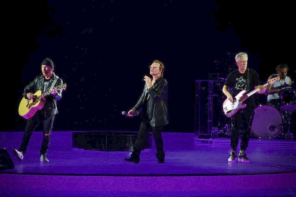 U2 en vivo en la Sphere de Las Vegas en octubre de 2023: The Edge con guitarra acústica amarilla, Bono cantando en el centro y Adam Clayton con bajo blanco, sobre un escenario iluminado de violeta (foto de archivo)