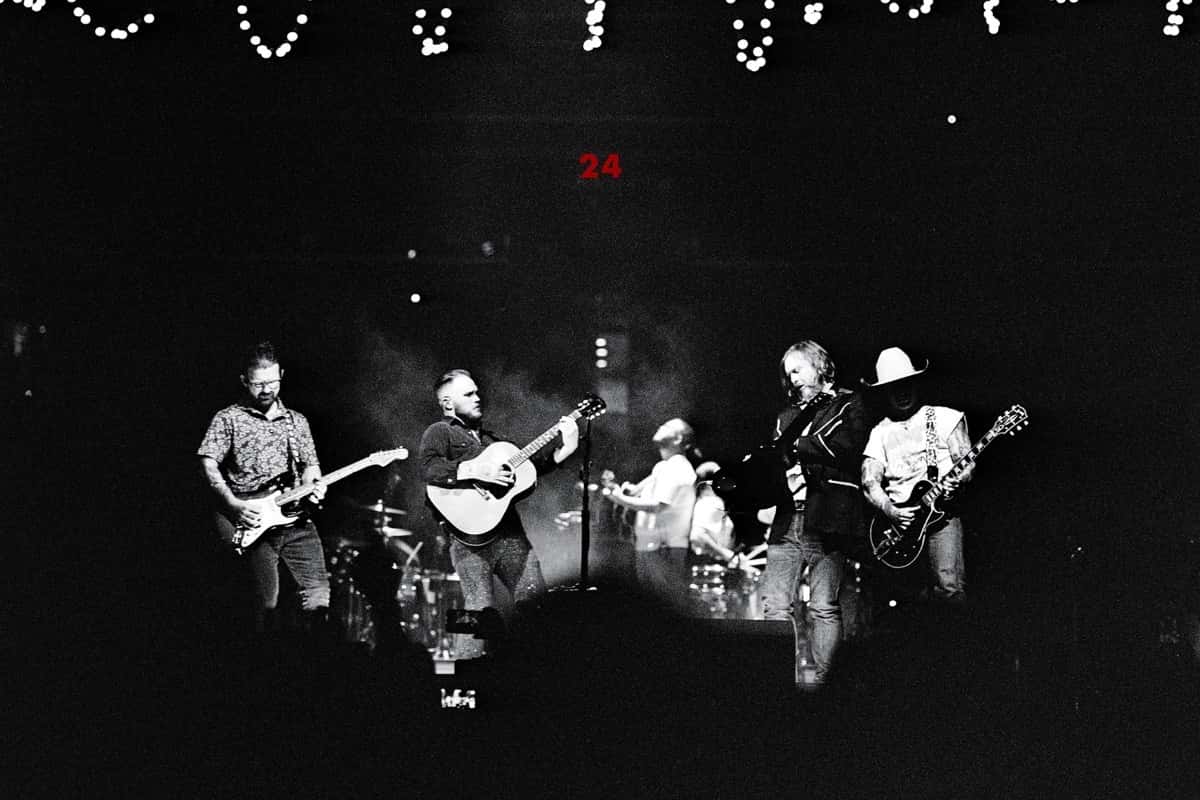 Imagen oficial de archivo de la web de Zach Bryan: foto en blanco y negro de Bryan y su banda en vivo, con guitarras eléctricas y acústica, bajo guirnaldas de luces y un 24 en rojo al fondo del escenario