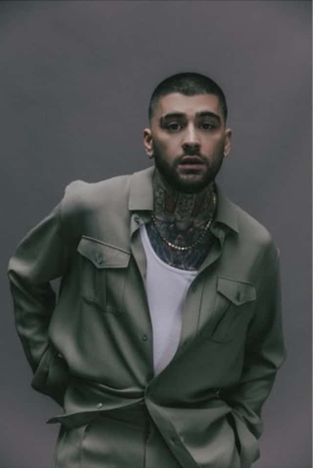 Foto de prensa de ZAYN de cuerpo casi entero, con el pelo rapado, barba y tatuajes en el cuello, camisa verde grisácea abierta sobre una musculosa blanca, cadena dorada y los brazos hacia atrás, frente a un fondo gris