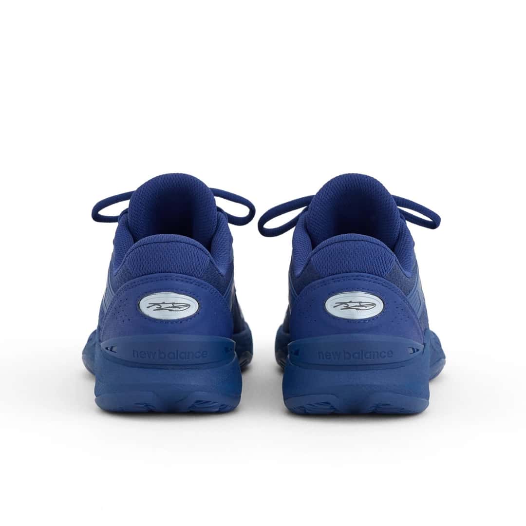 Vista trasera del par de New Balance Kawhi 5 Blue Oyster, en azul profundo, con el óvalo celeste con el logo de Kawhi en el talón, la palabra new balance grabada en la mediasuela y cordones azules