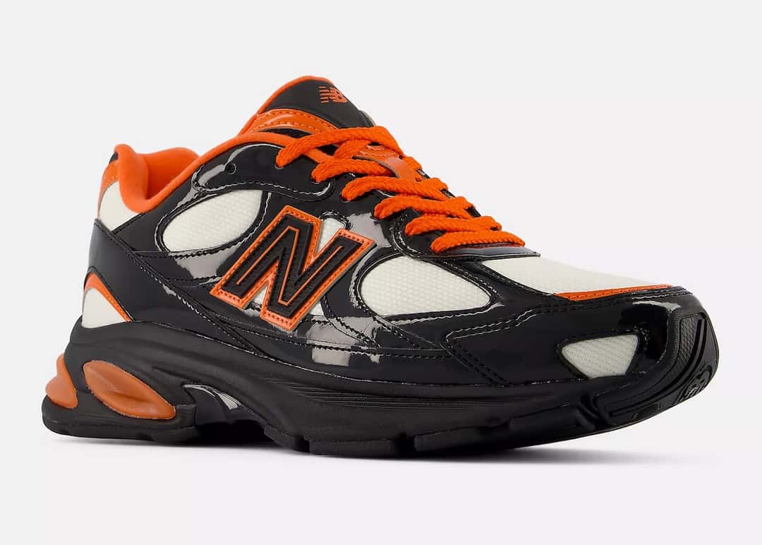 New Balance 2010 Halloween en vista tres cuartos sobre fondo gris claro, con la puntera de charol negro en primer plano, malla crema en la punta y el medio pie, logo N con borde naranja, cordones naranjas y pods naranjas en el talón