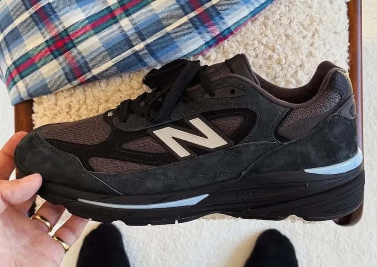 New Balance 990v7 Black sostenido en la mano de perfil, con gamuza negra, malla gris oscura, la N clara, la leyenda 990 en el talón y detalles celestes en la entresuela negra