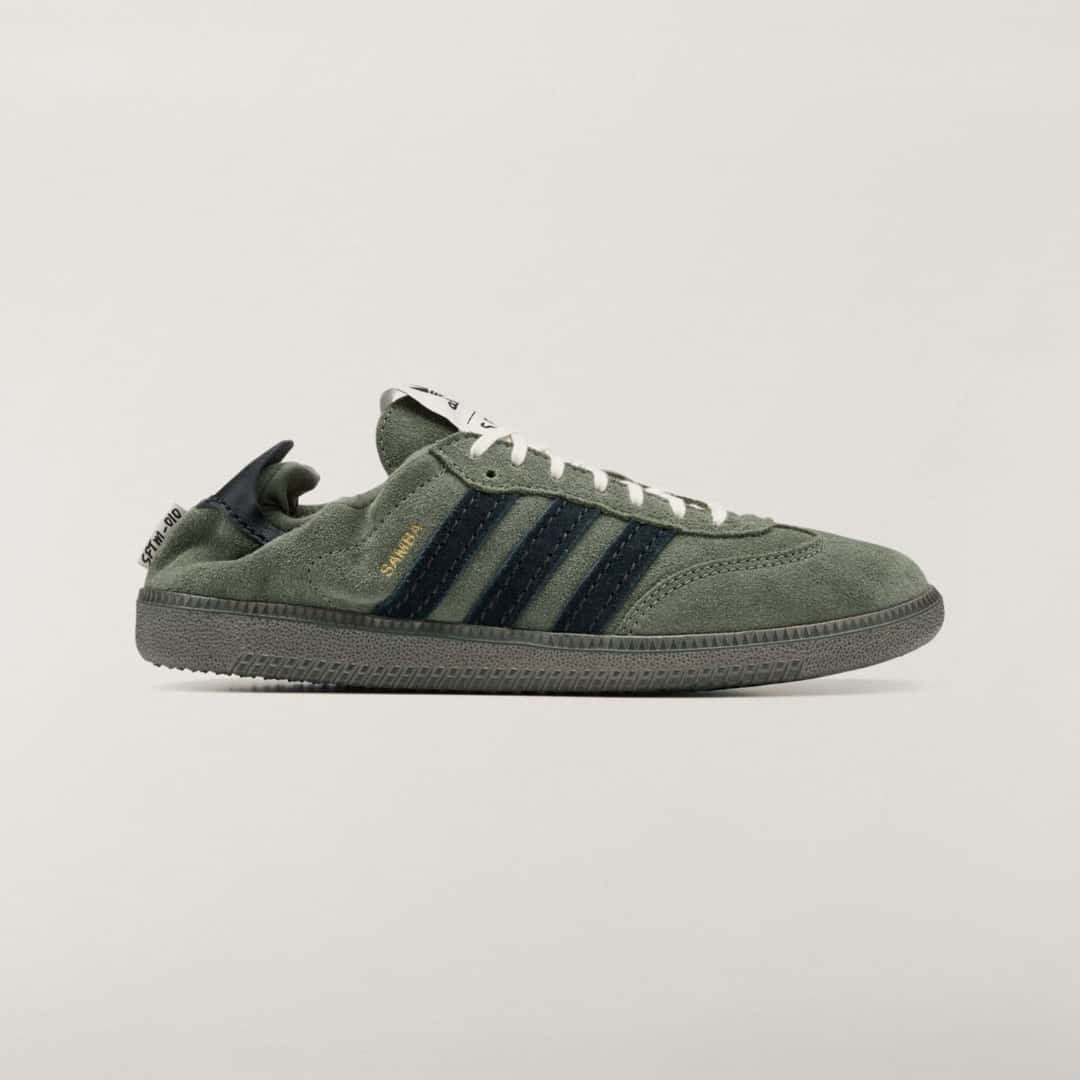 Vista lateral del adidas Samba OG Base Green de Song for the Mute en gamuza verde oliva, con las tres tiras en gamuza azul oscuro, la palabra Samba en dorado, cordones crema, suela verde grisácea y el talón doblado hacia adentro con la etiqueta SFTM-010