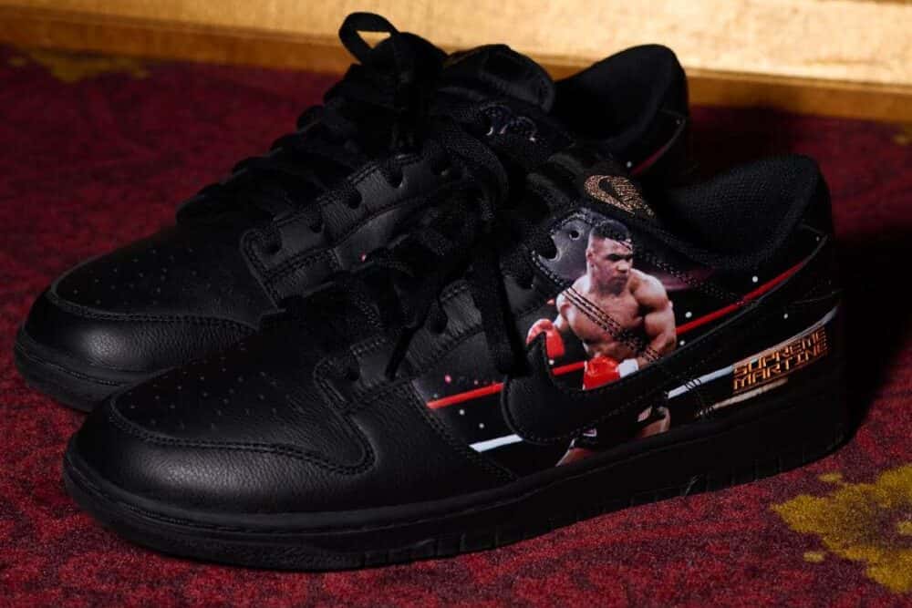 Par del Supreme x Martine Rose x Nike Dunk Low Tyson apoyado sobre una alfombra bordó, en cuero negro con cordones negros, la foto de Mike Tyson con guantes rojos sobre el lateral y las letras doradas Supreme Martine en el talón
