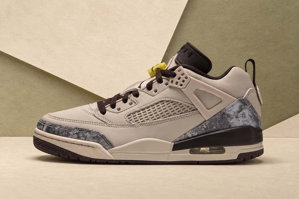 Jordan Spizike Low Realtree Camo de perfil sobre un fondo de planos crema y verde oliva, con upper de cuero crema, paneles de camuflaje gris en la punta y el talón, cordones marrón oscuro y traba de cordones amarilla