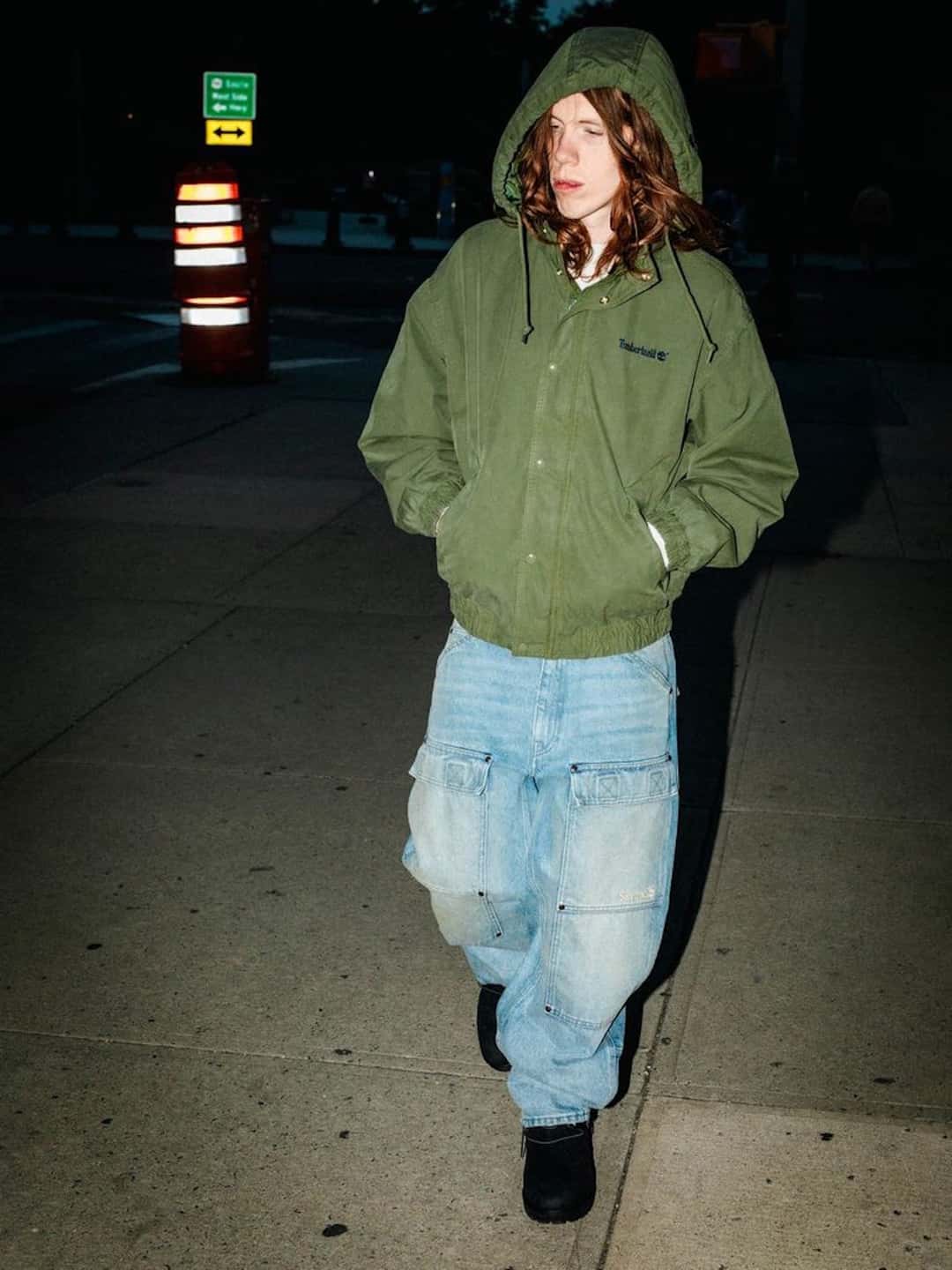 Modelo de noche en la calle con la Supreme x Timberland Ventile Utility Jacket verde oliva con capucha y jeans claros
