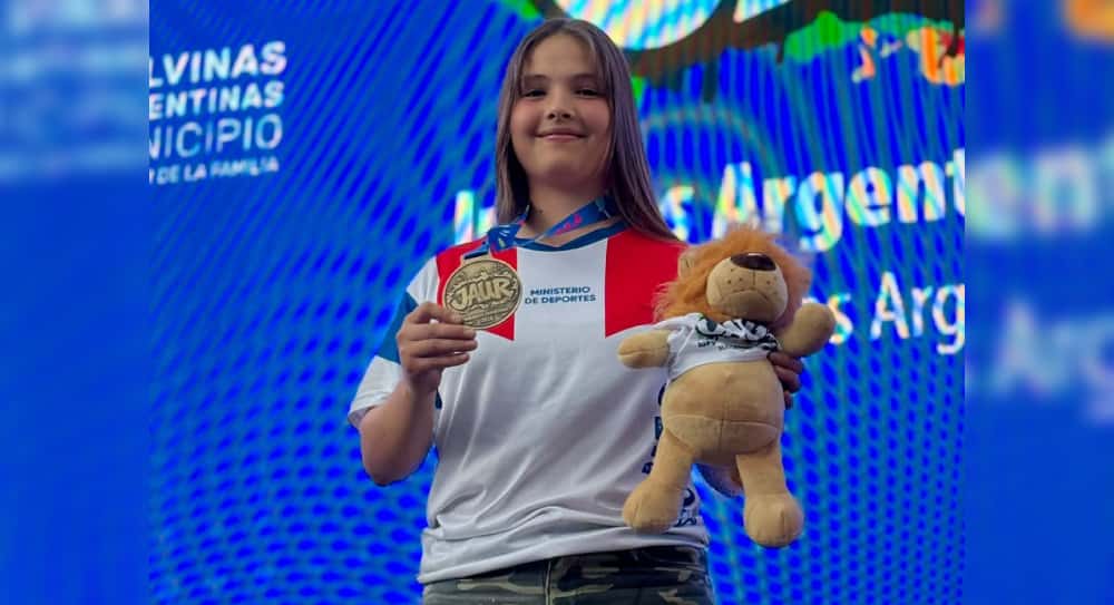 Ruth Feldick, con la camiseta de Misiones, muestra la medalla de oro de los Juegos Argentinos Urbanos y el peluche de la mascota frente a una pantalla con el logo de los JAUR