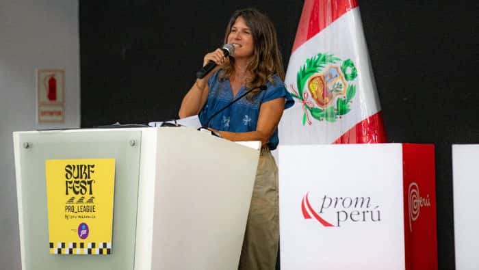 Sofía Mulanovich habla con un micrófono en un atril con el afiche amarillo del Surf Fest Pro League, junto a la bandera de Perú y un cartel de Promperú