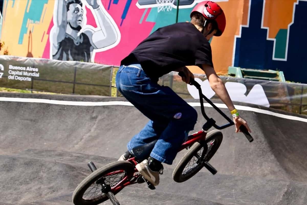 Un rider de BMX con casco rojo toma una curva peraltada del pumptrack del Parque Urbano del Parque Sarmiento, con murales de colores detrás