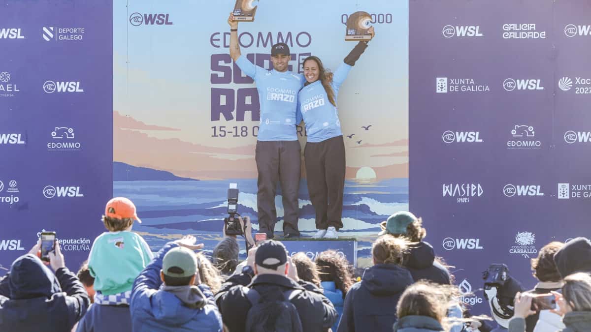 Yago Domínguez y Ariane Ochoa, con lycras celestes, levantan sus trofeos en el podio del Edommo Surf Pro Razo, delante de un cartel violeta con logos de la WSL y del público que les saca fotos