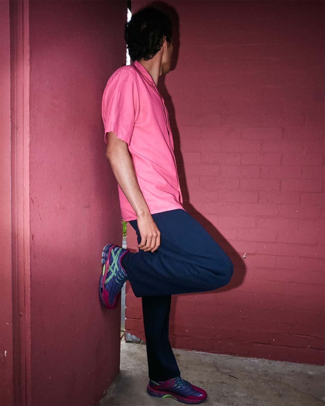 Modelo con camisa rosa y pantalón azul marino apoyado en una pared bordó, con una pierna flexionada contra el muro y la ASICS US8-S GEL-SJ33 Deep Plum/Fresh Grass puesta