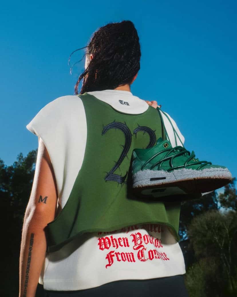 Foto de archivo: campaña del Air Jordan 3 x Teyana Taylor Concrete Rose: una mujer de espaldas con musculosa verde y el número 23 con espinas bordado, que lleva el par verde colgado del hombro bajo un cielo azul