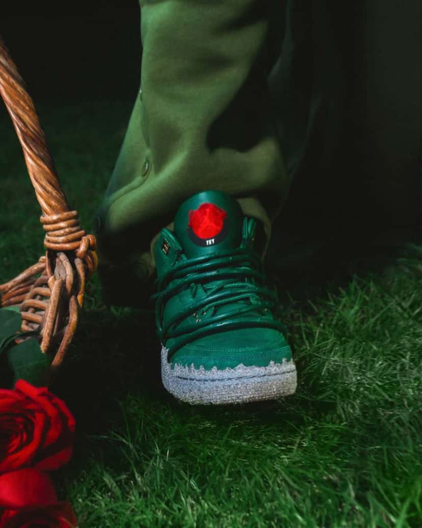 Foto de archivo: detalle del Air Jordan 3 x Teyana Taylor Concrete Rose sobre el pasto, con la rosa roja y la inscripción TEY en la lengüeta, los tallos verdes sobre el empeine y la mediasuela gris tipo concreto, junto a una canasta de mimbre y rosas rojas