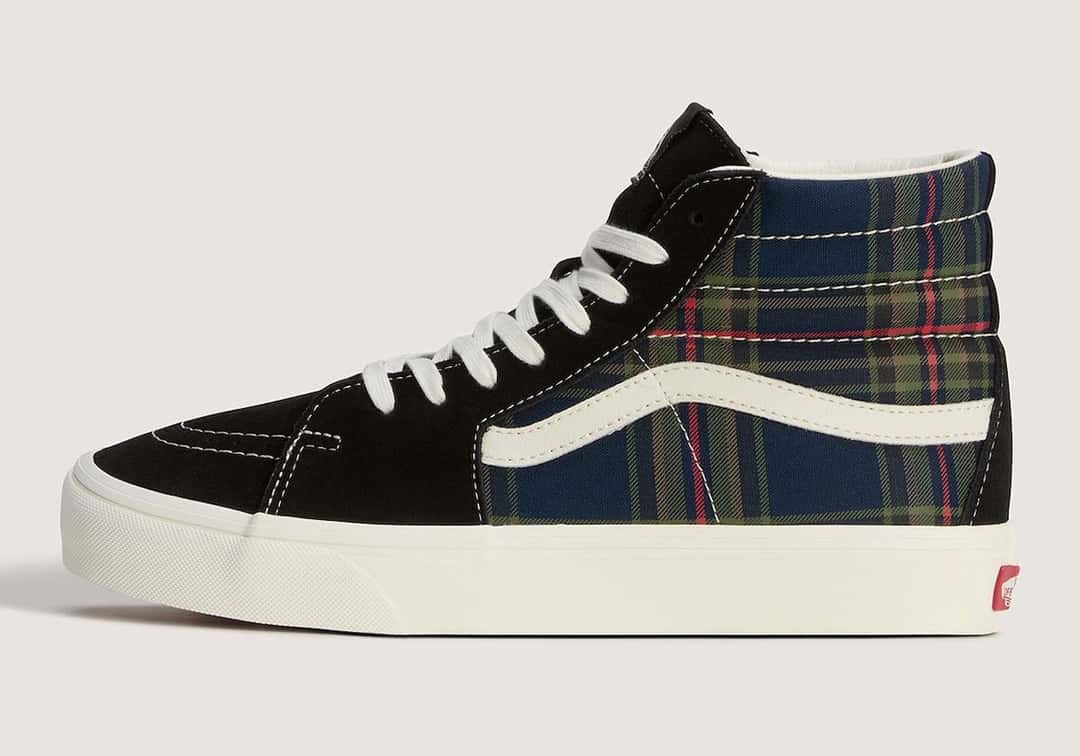 Vans Sk8-Hi Plaid Black/Multi de perfil sobre fondo crema, con gamuza negra en la punta y el empeine, caña de cuadros azul marino con líneas verdes y rojas, Sidestripe crema, cordones blancos, costuras blancas a la vista y suela crema con la etiqueta roja de Vans en el talón