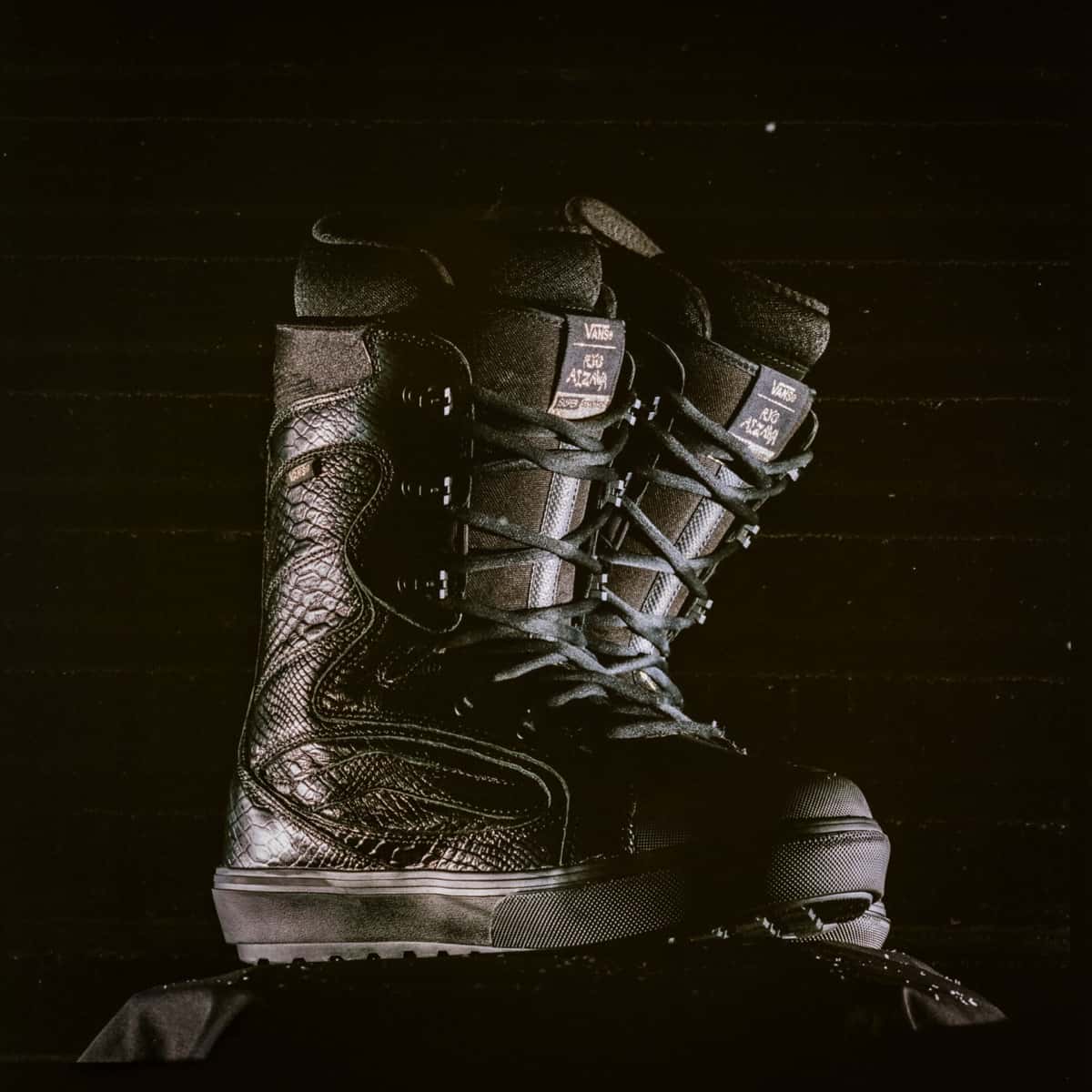Par de botas de snowboard Vans Super Standard negras con textura de serpiente, cordones negros y lengüeta con la firma de Ryo Aizawa, fotografiadas sobre fondo oscuro