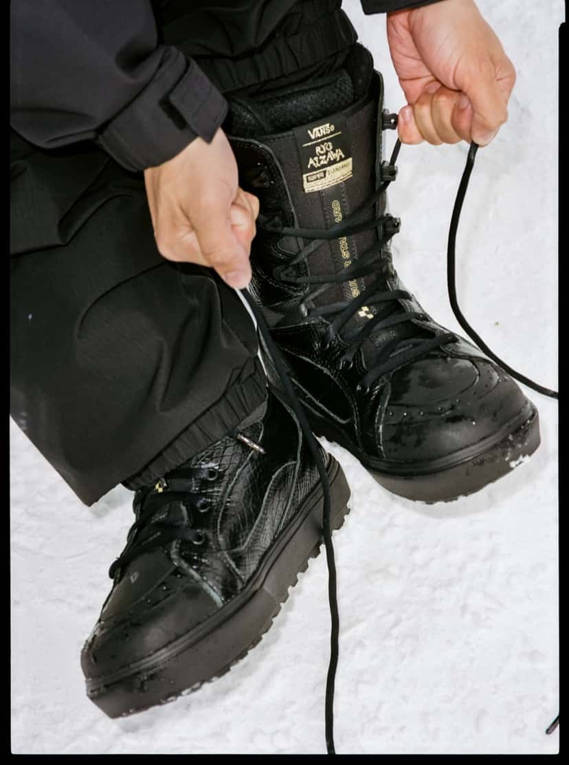 Rider atándose los cordones de las botas Vans Super Standard negras con textura de serpiente sobre la nieve; en la lengüeta se lee la firma de Ryo Aizawa
